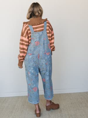 Shot from behind, she's wearing floral-printed denim overalls over a striped turtleneck sweater — the kind of layered, lived-in outfit that looks like it came together effortlessly but was absolutely thought through.