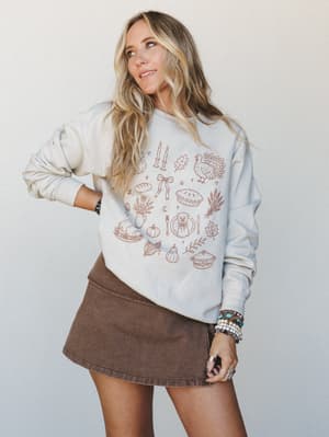 A relaxed cream pullover sweatshirt printed with a warm collection of Thanksgiving-themed line art icons — turkey, pie, pumpkins, wheat, candles — styled with a chocolate brown mini skirt and layered boho jewelry for a cozy-meets-cute fall look.