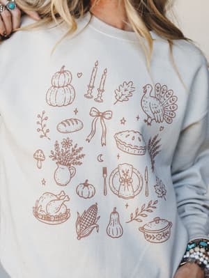 A close-up of a sand-colored crewneck sweatshirt covered in a warm, illustrated Thanksgiving motif — think pumpkins, turkey, pie, candles, and corn all sketched in a rusty terracotta line-art style that feels like your coziest autumn daydream.