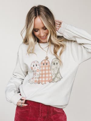 A cream pullover sweatshirt with a trio of boho-patterned ghost graphic — floral, gingham, and daisy-print — styled with red corduroy pants and layered gold necklaces, giving off the coziest not-too-spooky Halloween energy.