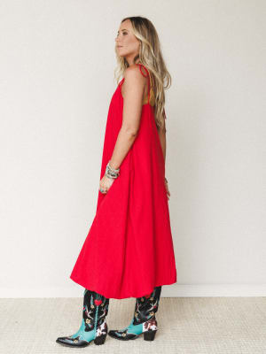 A flowy red maxi bubble dress with tie shoulders and a relaxed silhouette, styled with bold embroidered cowboy boots and stacked turquoise bracelets — shot from the side against a soft white studio wall.