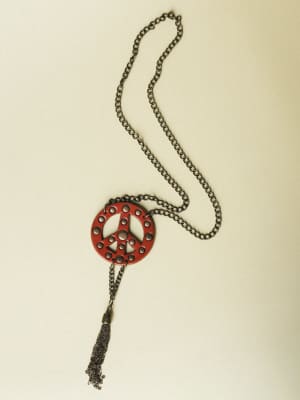 A long chain necklace laid flat against a neutral background, featuring a deep red leather peace sign pendant with circular cutouts and a dangling chain tassel — the kind of piece that does all the talking.