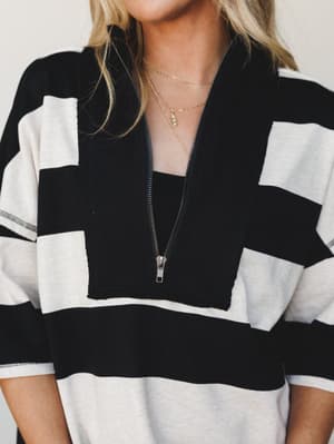 A close-up of a bold black and cream wide-stripe half-zip top styled with layered gold chain necklaces — relaxed, confident, and just the right amount of casual-cool.