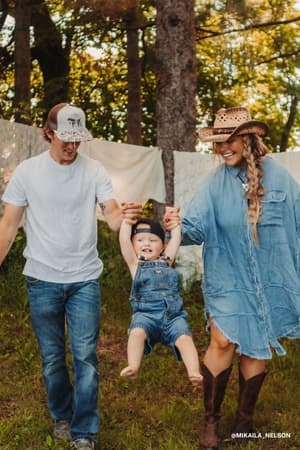 A man and woman in casual wear swing a smiling toddler outdoors, with trees and white sheets behind them—capturing a carefree vibe ideal for the Ophelia Oversized Denim Dress - Washed Denim.