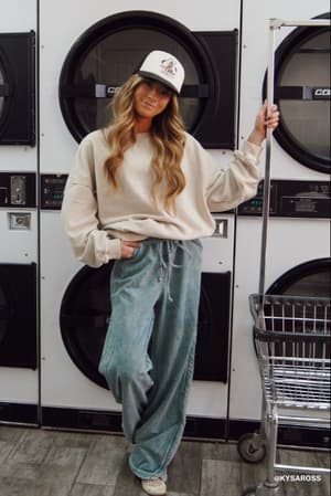 A woman in a cream sweatshirt, So Comfy Wide Leg Pant Full Length - Teal, and a white cap leans against a cart in a laundromat, with large washers and dryers visible behind her.