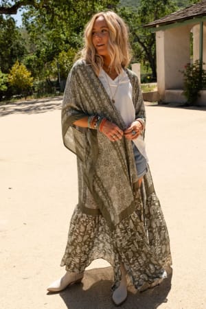 A woman stands outdoors wearing the Paisley Tapestry Free Flow Duster Kimono – Olive over a white top and denim shorts, accessorized with bracelets, rings, and white boots. Her wavy blonde hair complements the casual look. Trees and a building are in the background.