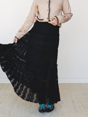 A black lace tiered maxi skirt caught mid-swirl, paired with a camel cardigan with black blanket-stitch trim and turquoise cowboy boots — the kind of outfit that has a whole personality.