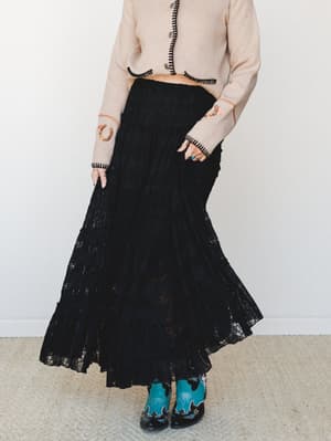 A black tiered lace maxi skirt paired with a cream cropped cardigan featuring black whipstitch trim and embroidered sleeves, finished with teal western cowboy boots — the kind of outfit that feels like it has a whole personality.