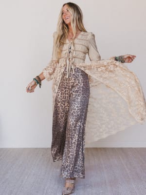 Woman wearing a beige top and leopard print pants with a flowing fabric in a minimal indoor setting.