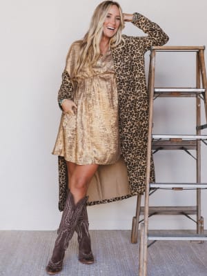Woman wearing a gold sequin dress and leopard print cardigan with brown cowboy boots, standing next to a ladder.