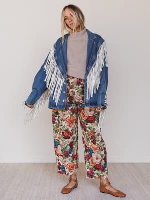 Person wearing a denim jacket with white fringe and floral pants on a plain background