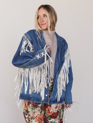 Woman wearing a blue denim jacket with white fringe against a plain background