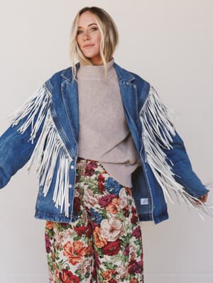 Person wearing a blue denim jacket with white fringe, gray sweater, and floral pants on a plain background