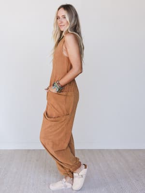 Side view of a woman wearing a rust sleeveless jumpsuit against a white background