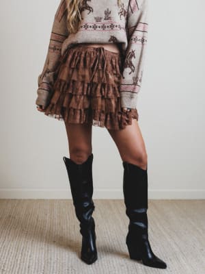 Person wearing a patterned sweater, brown ruffled skirt, and black knee-high boots on a plain background