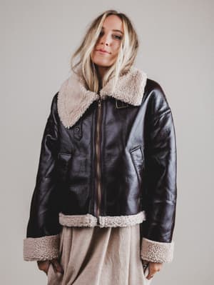 Person wearing a black leather jacket with beige shearling collar and cuffs on a plain background