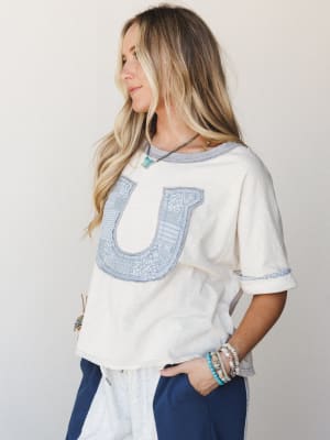 A relaxed cream tee with a patchwork horseshoe appliqué in soft blue takes center stage — styled with navy pants and stacked boho bracelets for that effortless, personality-first vibe.