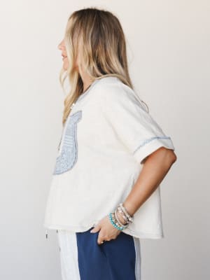 A relaxed, side-profile shot of a cream oversized tee with a dusty blue embroidered bib-style chest panel and rolled sleeves, styled with navy wide-leg pants and a stack of beaded bracelets — the kind of effortless outfit that looks like it came toge