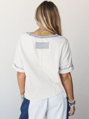 A back-facing shot of a relaxed cream tee with contrast periwinkle trim and a visible patch detail — styled with wide-leg pants and stacked bracelets for that effortless, lived-in feel.