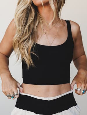 A close-up torso shot of a black cropped bralette styled with layered gold necklaces, stacked silver and turquoise rings, and a glimpse of a cream ribbed waistband below — effortless and a little collected, like she got dressed without trying too har