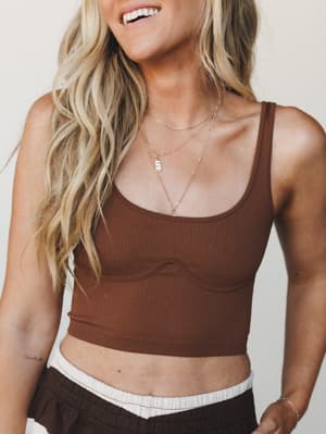 A close-up lifestyle shot of a ribbed brown crop bralette styled with wide-leg brown pants and layered gold necklaces — effortless and warm, like a Saturday that has nowhere to be.