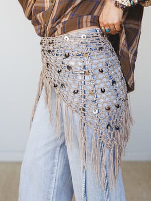 A close-up of a macramé fringe hip belt layered over wide-leg light wash jeans and a plaid shirt, styled with stacked boho rings and bracelets — the kind of piece that makes a whole outfit feel intentional.
