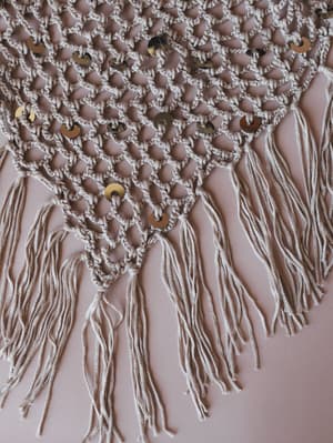A close-up detail shot of an open-weave crochet belt or waist accessory in warm oatmeal, scattered with gold coin sequins and finished with cascading fringe tassels — laid flat against a blush background.