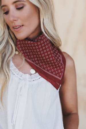 A woman with long blonde hair and a gold necklace wears a sleeveless white blouse and the Rylee Flower Field Bandana Scarf in rust, standing against a light, blurred background.