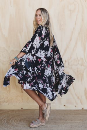 A woman with long blonde hair smiles as she twirls in front of a light wooden wall, wearing the Free Flowin' Butterfly Sleeve Kimono in black.