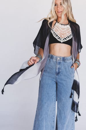 A woman with long blonde hair wears high-waisted wide-leg jeans, a black Crochet High Neck Bralette, layered bracelets, and a sheer black-and-white shawl, standing against a plain white background.