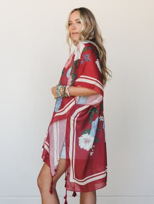 A flowy floral kimono in deep crimson with cream border stripes and tassel hem styled over denim shorts — the kind of layering piece that makes a casual outfit feel intentional.