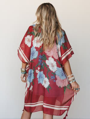 A flowy floral kimono in deep rust red with oversized blooms in dusty blue, blush, and white — shot from the back, styled with stacked bracelets and a relaxed, effortless feel.