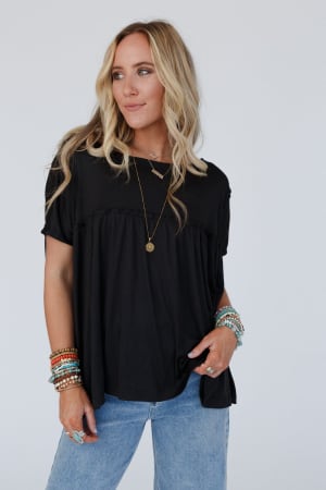 A woman with long blonde hair wears the Sunday Morning Babydoll Top - Black, styled with blue jeans, layered necklaces, and colorful bracelets, standing against a plain light background and looking slightly to the side.