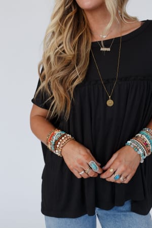 A woman with wavy blonde hair wears the Sunday Morning Babydoll Top - Black, paired with blue jeans, layered necklaces, beaded bracelets, and turquoise rings. Her face is partially out of frame.