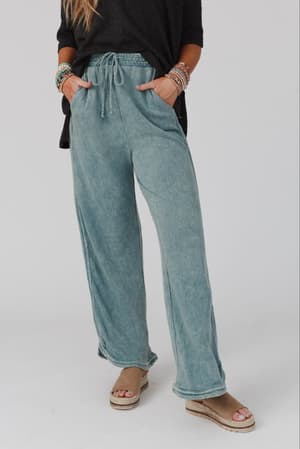 Wearing the So Comfy Wide Leg Pant Full Length in teal with a drawstring waist, a loose black top, beaded bracelets, and platform sandals, a person stands against a plain light background. Their face is not visible.