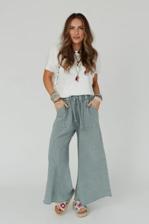A woman with long wavy hair stands against a white background, wearing a white t-shirt, the Relaxing Robin Wide Leg Pant in Faded Teal, colorful bracelets, and embroidered sandals, with her hands in her pockets.