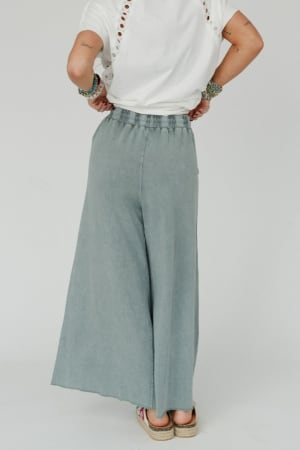 A person faces away, wearing a white eyelet short-sleeve top and the Relaxing Robin Wide Leg Pant in Faded Teal. They accessorize with bracelets and flat shoes for a comfortable, boho-inspired look.