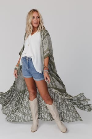 A woman with blonde, wavy hair poses against a plain light background wearing the Paisley Tapestry Free Flow Duster Kimono in Olive over a white blouse and denim shorts, styled with tan cowboy boots, bracelets, and necklaces.