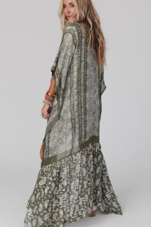 A woman in a green and white maxi dress and stacked bracelets stands sideways, looking over her shoulder, styled effortlessly with the Paisley Tapestry Free Flow Duster Kimono in Olive. She has long wavy blonde hair against a plain light background.