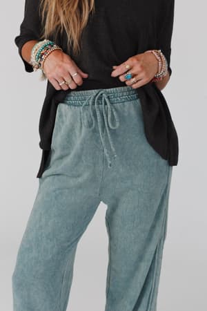 A person wearing a black long-sleeve shirt, teal So Comfy Wide Leg Pant Full Length, and several colorful beaded bracelets and rings, with their hand resting on their hip.
