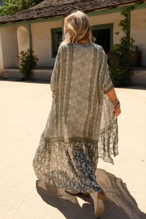 A woman with wavy blonde hair, seen from behind, wears the Paisley Tapestry Free Flow Duster Kimono in Olive and wedge sandals as she strolls outside near a rustic building with green-trimmed windows and flowering bushes.