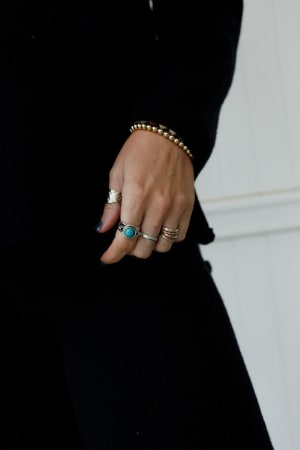 A close-up shows a hand wearing several rings, including the Kapalua Bay Adjustable Turquoise Ring - Silver, and a beaded bracelet. The dark outfit and blurred light background highlight the boho jewelry accessories.