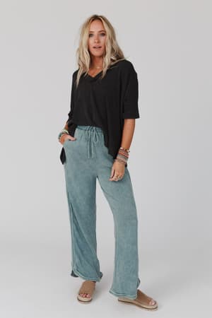 A woman with long blonde hair wears a loose black top, the So Comfy Wide Leg Pant Full Length in teal, beige sandals, and colorful bracelets, standing against a plain white background.