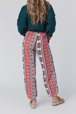 A person with long wavy hair, in a dark green sweater and The Nest Staycation Wide Leg Drawstring Pant - Red Clay, stands facing away. They wear beige slip-on shoes against a plain, light background.