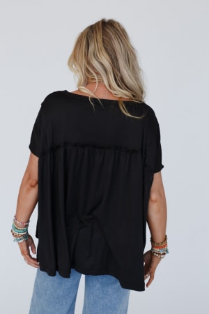 A person with long blonde hair, facing away from the camera, wears the Sunday Morning Babydoll Top in black with light blue jeans and colorful beaded bracelets. The background is plain white.