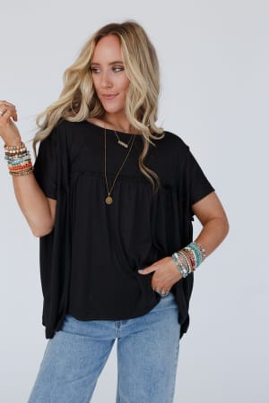 Woman with long blonde hair wears the Sunday Morning Babydoll Top in Black, styled with light blue jeans, layered necklaces, and colorful beaded bracelets, posing with one hand raised against a plain light background.