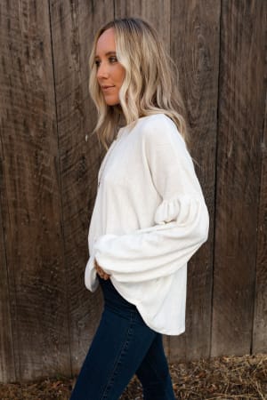 A woman with long blonde hair wears The Nest Sweet Heart Balloon Sleeve Knit Sweater in cream and dark jeans, standing sideways by a rustic wooden fence outdoors.