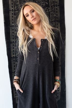 A woman with long blonde hair wears the Simply Comfort Jumpsuit - Dark Gray, styled with several bracelets, standing before a black patterned tapestry and gazing at the camera with a relaxed expression.