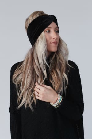 A woman with long, wavy blonde hair wears the Everyday Velvet Headband in Black and a black knit sweater. She has several beaded bracelets on her wrist and gazes slightly to the side against a plain light background.