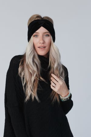 A woman with long, wavy blonde hair wears the Everyday Velvet Headband in Black and a black sweater. She holds a lock of her hair, displaying several beaded bracelets, and stands against a plain light background.
